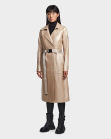 A woman faces forward against a plain light background, wearing the Rudsak KIRALY - 8123553 Metallic Ivory trench coat with a belt, paired with a black turtleneck, leggings, and combat boots.