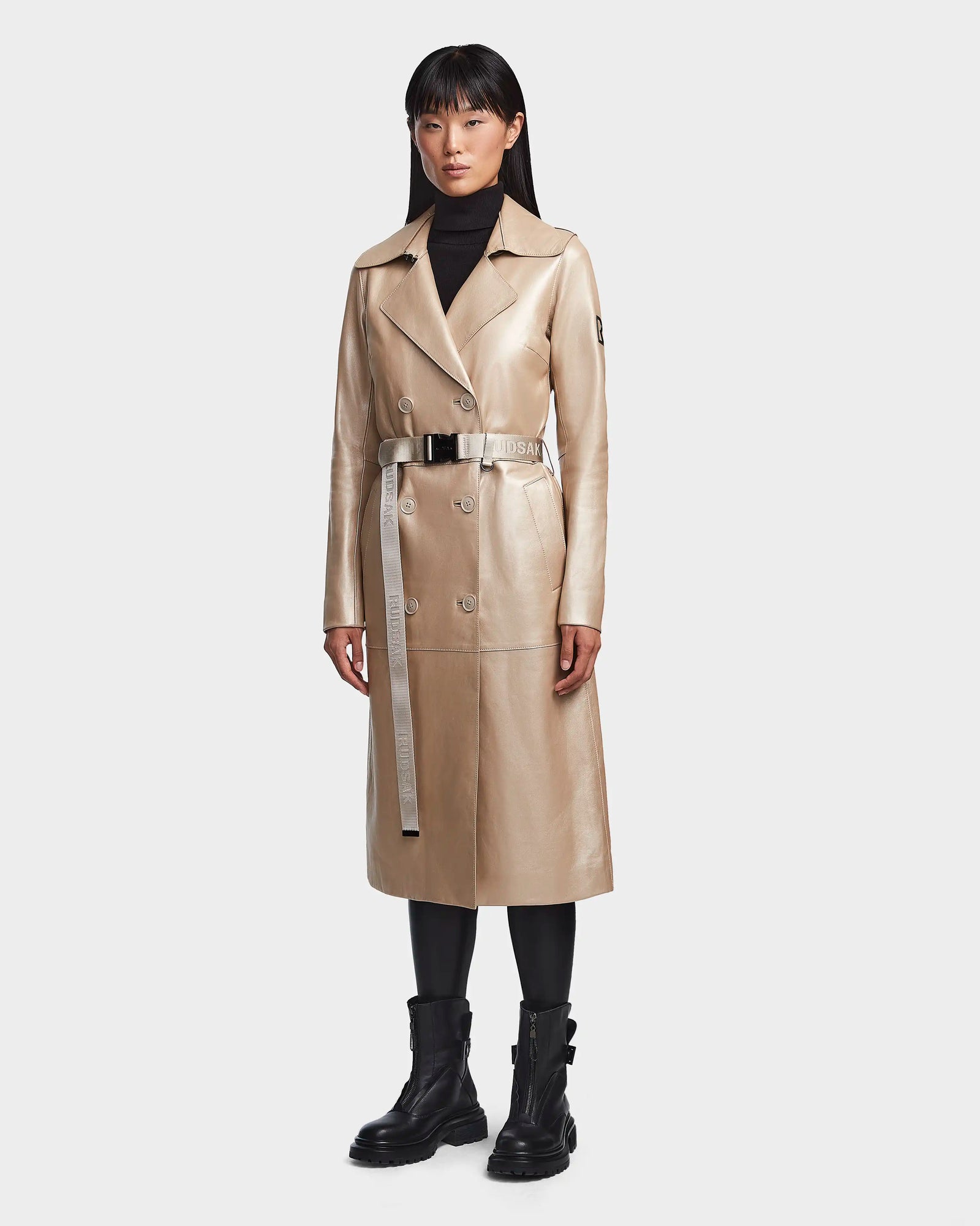 A woman faces forward against a plain light background, wearing the Rudsak KIRALY - 8123553 Metallic Ivory trench coat with a belt, paired with a black turtleneck, leggings, and combat boots.