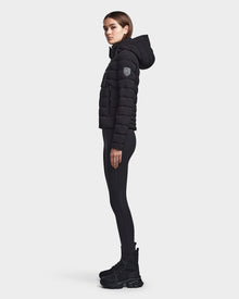 A woman stands in profile wearing the RUDSAK MELISSA - 8122551 BLACK puffer jacket with a hood, paired with black leggings and boots, against a plain light gray background.