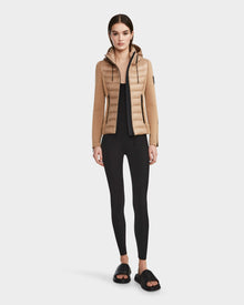 A woman faces forward in a RUDSAK MABS - 8122020 TOFFEE women’s puffer jacket with a hood, paired with black leggings and black slide sandals. She has straight brown hair pulled back, a neutral expression, and stands against a plain white background.