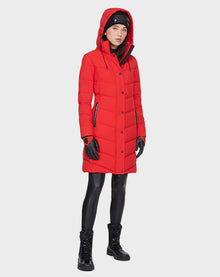 A woman stands against a plain light gray background wearing the Rudsak ELISA - L121512 POPPY RED quilted down puffer, black gloves, leggings, lace-up boots, and a beanie.
