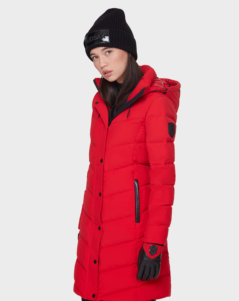 A woman in the Rudsak ELISA - L121512 POPPY RED quilted down puffer, with black gloves and a black knit beanie, stands against a plain white background, gazing slightly to the side.