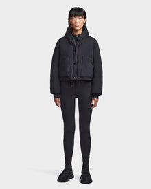 A person with straight black hair faces forward, wearing the Rudsak BALA - 8123614 Orange sustainable puffer women’s winter coat, black leggings, and black shoes against a plain white background.