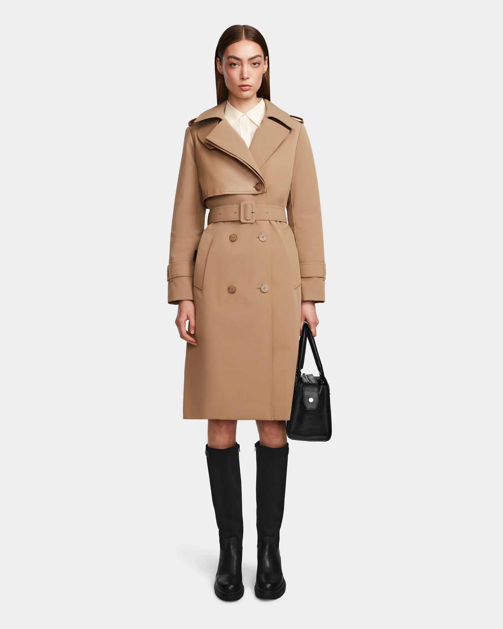 A woman with straight brown hair and a neutral expression wears the Rudsak KINDA - 8124690 Camel trench coat, paired with a white collared shirt, black knee-high boots, and holds a black handbag.