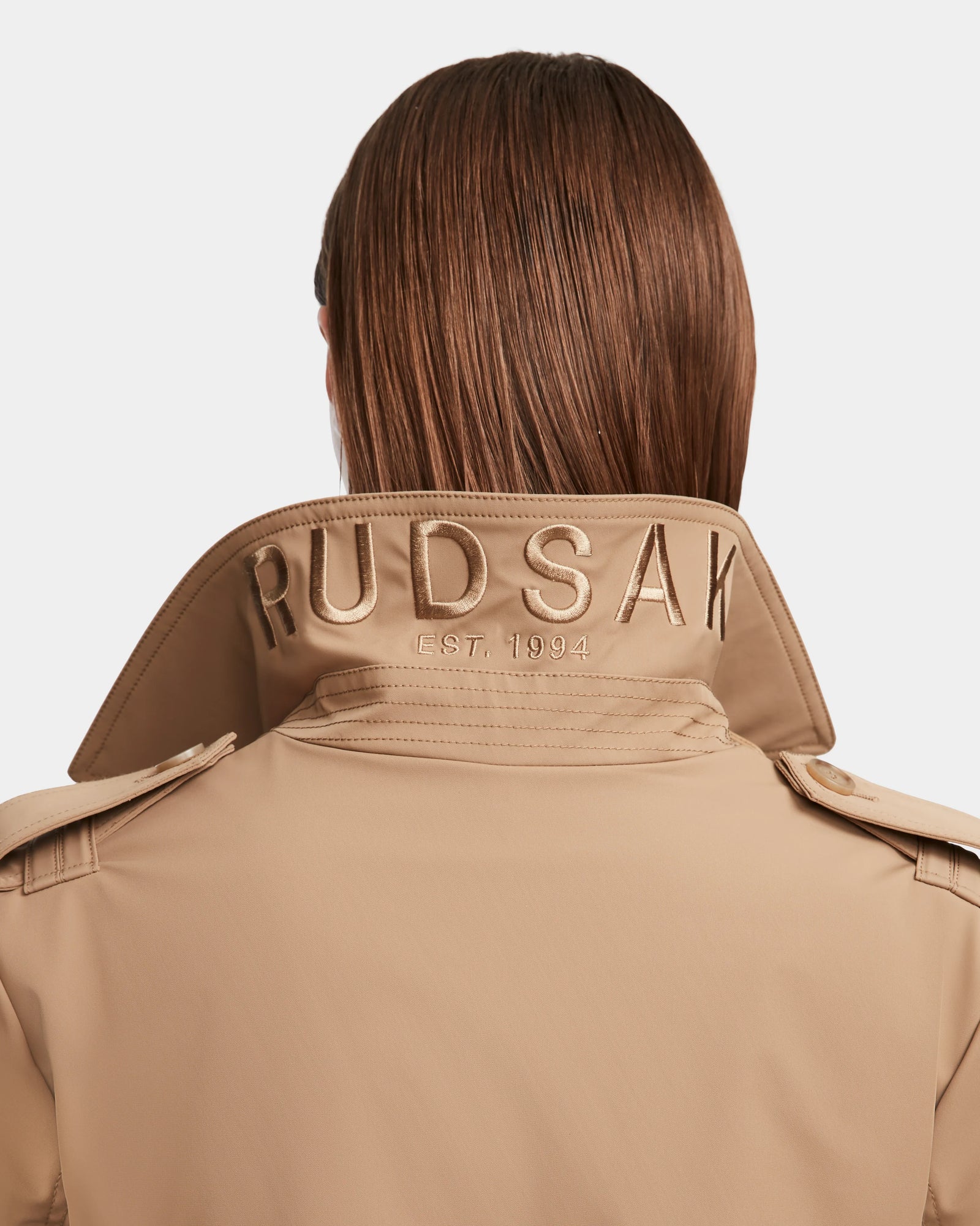 A person with straight brown hair is shown from behind, wearing the KINDA - 8124690 Camel womens trench coat by Rudsak, featuring RUDSAK EST. 1994 embroidered on its upturned collar.