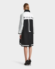 A woman with straight, shoulder-length hair and black heels faces away, wearing the Rudsak MIRELLE - 8124055 Black coat featuring a tailored silhouette and logo on the back. The background is plain light gray.