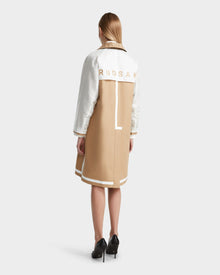 A woman with straight, blonde hair models the Rudsak MIRELLE - 8124055 Khaki womens trench coat with a tailored silhouette and RUDSAK on the back, wearing black heels and facing away against a plain background.