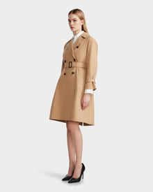 A woman models the Rudsak MIRELLE - 8124055 Khaki trench, featuring a belted, knee-length tailored silhouette with black buttons, worn over a white shirt and paired with black heels against a plain light background.