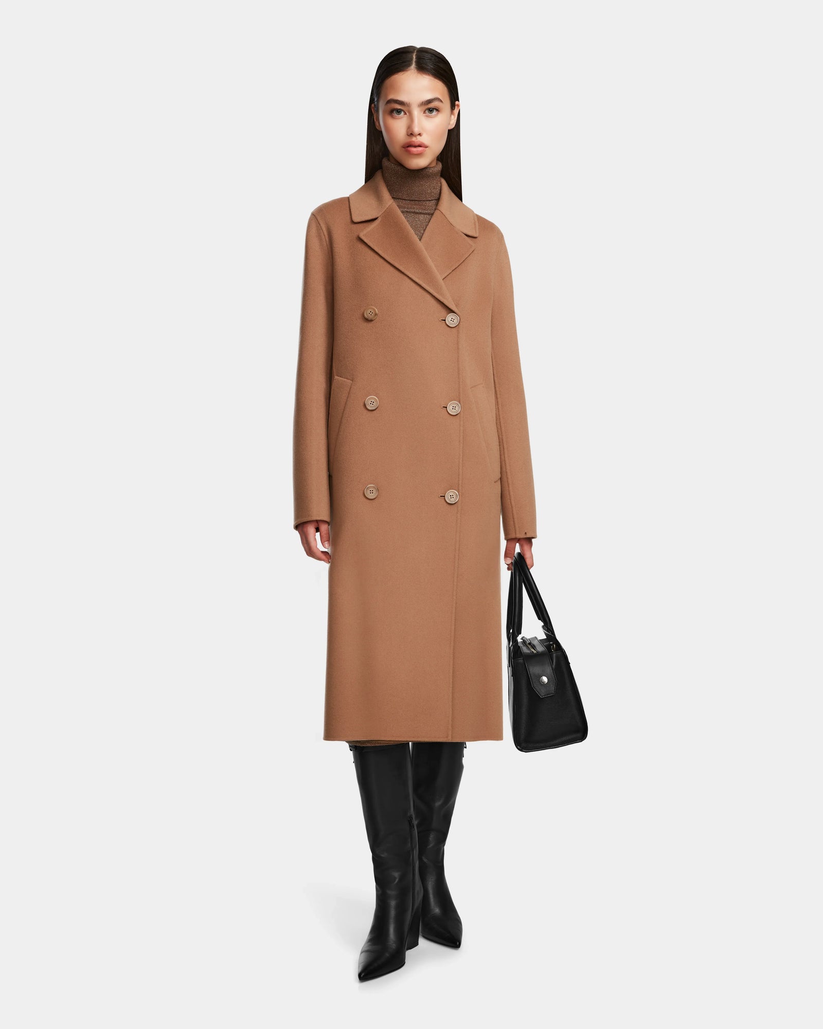 A woman with straight dark hair and a neutral expression stands against a plain background wearing the Rudsak LAYLAH - 8124699 knee-length camel coat, styled with black knee-high boots and a black handbag.