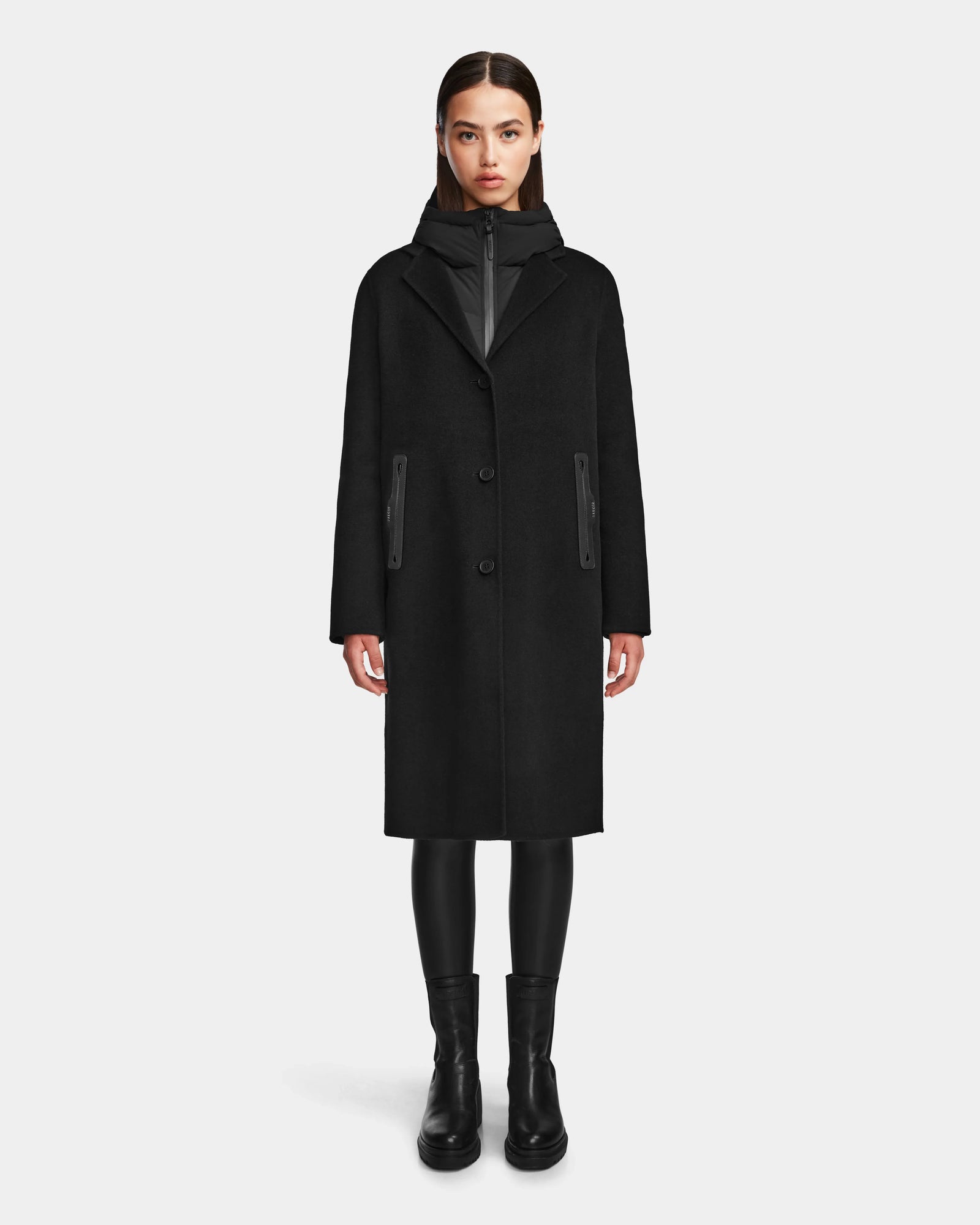 A woman faces forward against a white background, wearing Rudsak’s ARLA - 8124579 Black wool coat with leather sleeves, paired with black leggings, a black zip-up underneath, and black boots.