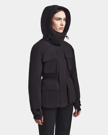 A woman in the Rudsak LUX - 8123618 Black women’s ski jacket with RECCO technology and large front pockets stands against a light gray background, gazing slightly to the side.