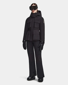 A person stands facing forward against a plain white background, dressed in the Rudsak LUX - 8123618 Black women’s ski jacket, paired with matching ski pants, gloves, boots, and large ski goggles for a sleek winter look.