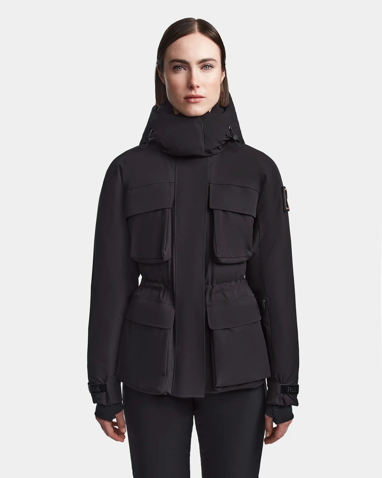 A woman with straight brown hair models the Rudsak LUX - 8123618 Black ski jacket, featuring four large front pockets, against a white background—a chic choice for winter style.