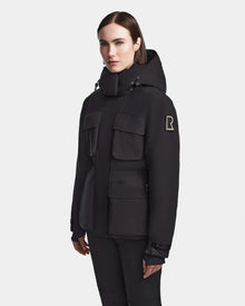 A woman with straight brown hair wears the Rudsak LUX - 8123618 Black womens ski jacket, featuring multiple front pockets, RECCO technology, and an upper arm R patch, standing against a plain light background.
