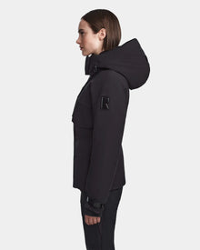 A woman in profile, facing left, models the Rudsak LUX - 8123618 Black women’s ski jacket with a prominent R patch on the upper sleeve. A white background accentuates her chic winter style.