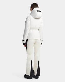 A person stands facing away, wearing the Rudsak LUX - 8123618 Iceland white waterproof ski jacket with RECCO technology and matching white pants featuring black panels and flared legs. They complete the sporty look with black shoes on a light gray background.