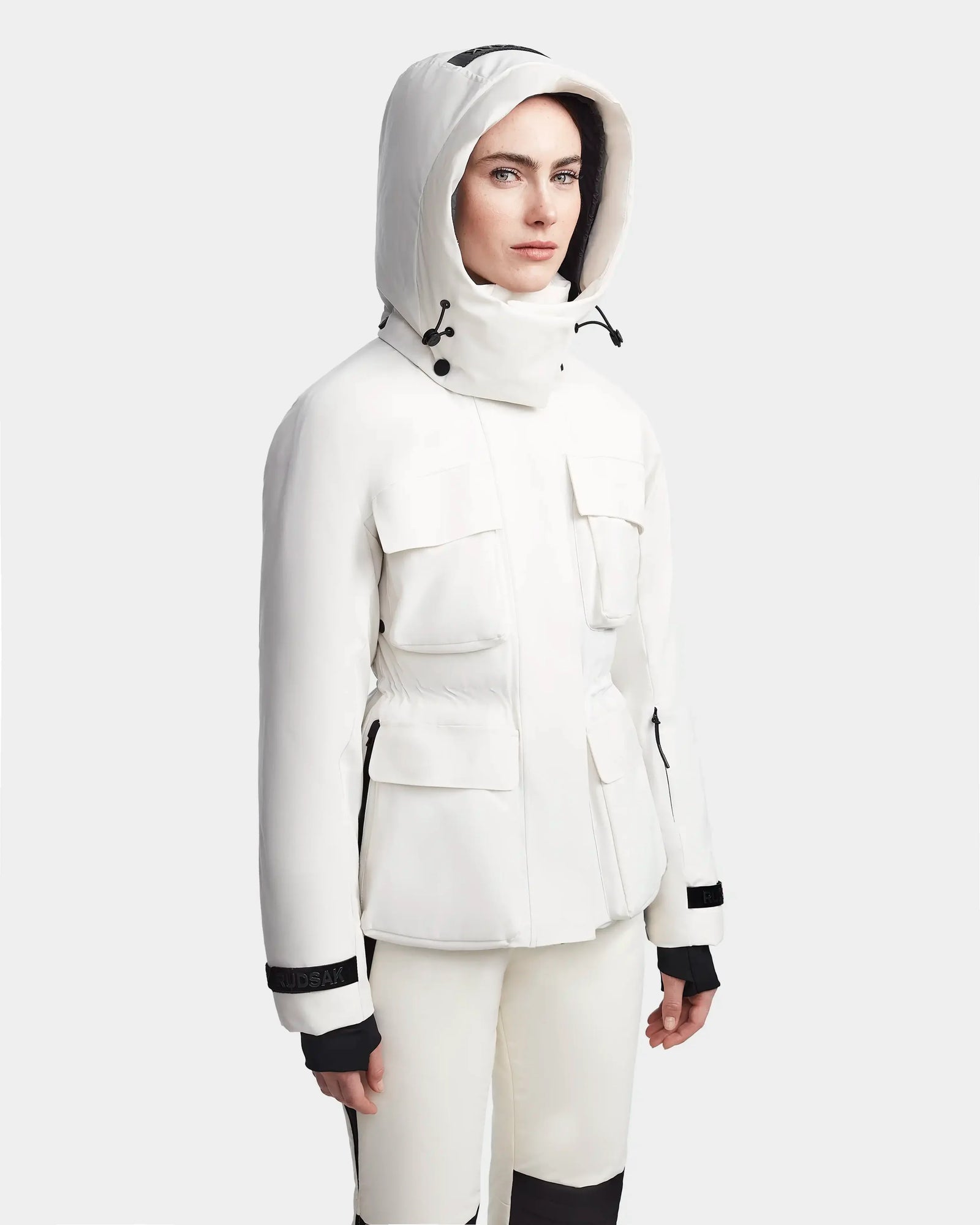 A person wearing the Rudsak LUX - 8123618 Iceland white women’s ski jacket with black accents, multiple pockets, and RECCO technology stands against a plain light gray background, highlighting functional winter style.