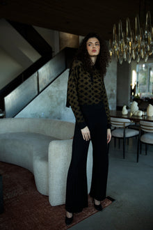 Curly-haired brunette model standing in a living room, wearing the golden black Amelie Polo styled with a French tuck and black alpaca silk stretch trousers.
