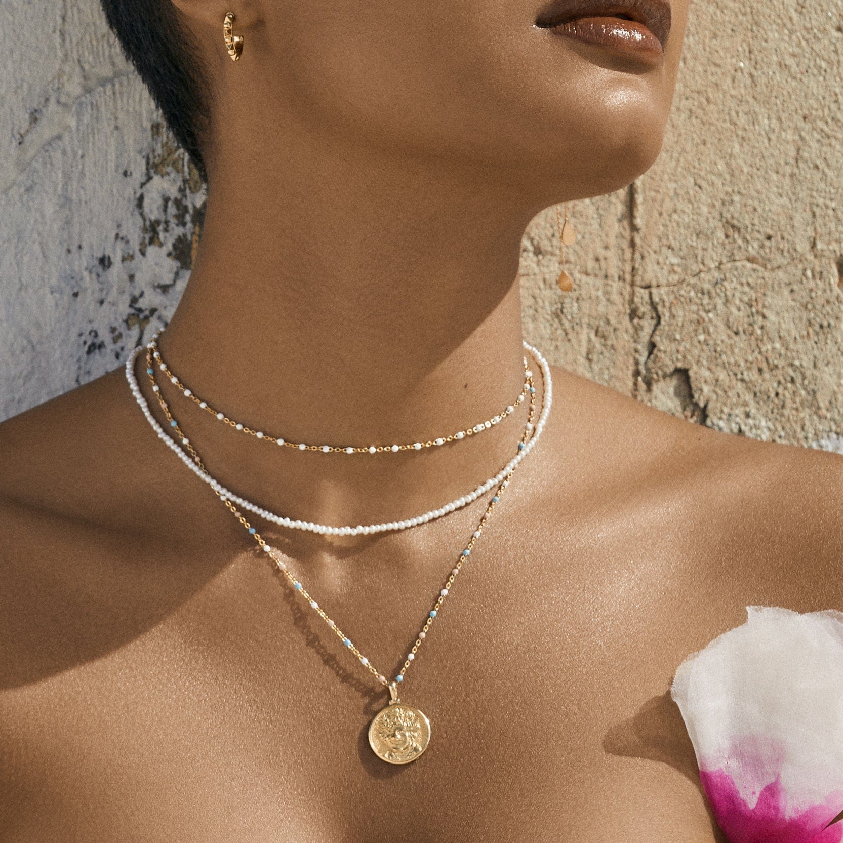 Close-up of a person wearing layered necklaces, including gold chains, a medallion, and the Trans Pride Beaded Enamel Necklace by Awe Inspired, against a textured wall with an off-the-shoulder top featuring a pink and white flower detail.