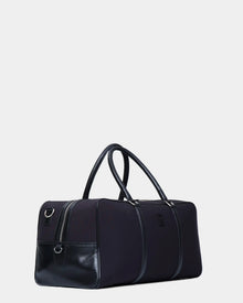 The Rudsak OLIVER - 9323507 Black unisex duffel bag, featuring leather accents, double handles, and a zippered front pocket, is displayed against a light gray background—ideal for luxury travel on any occasion.