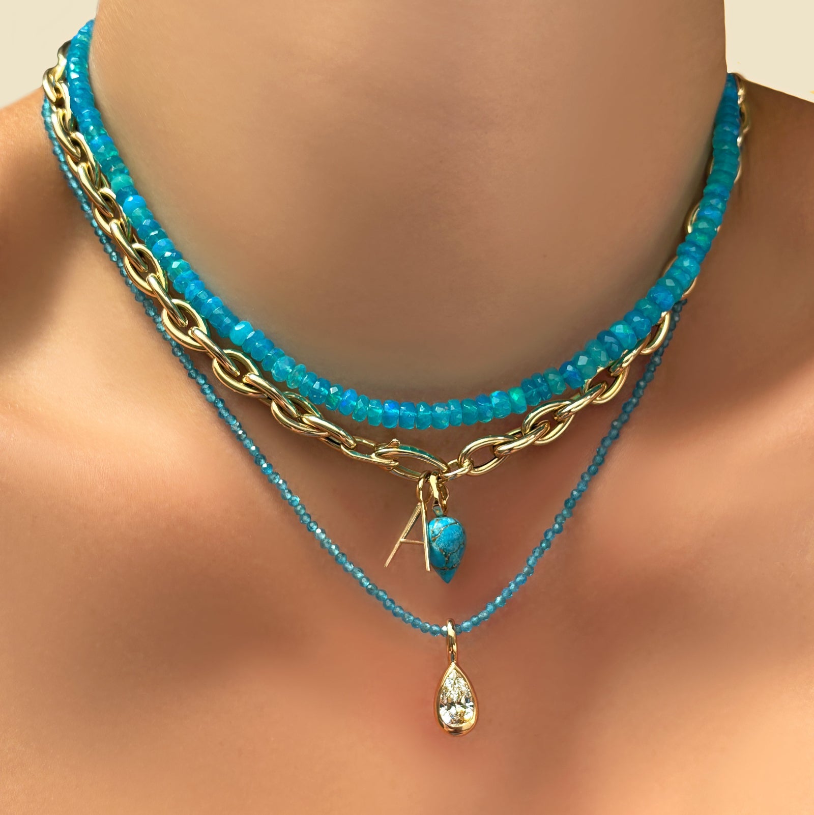 bright blue faceted opal necklace with  drop chain necklace, letter A charm, turquoise drop charm, blue apatite slim gemstone necklace, and molten pear solitaire charm on a model's neck.
