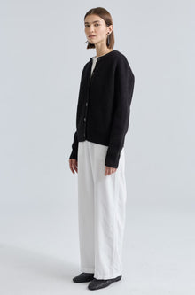 Person wearing a black cardigan, white pants, and dark shoes on a light gray background