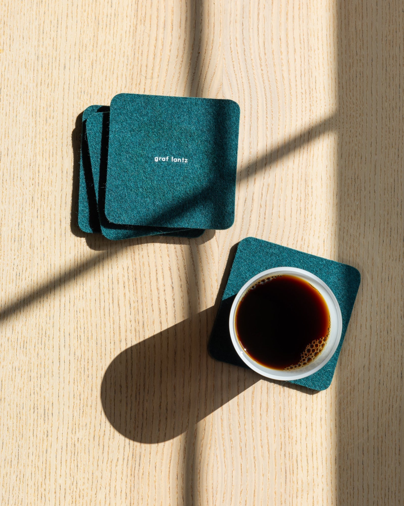 Four square coasters in color Kombu on a wooden tabletop, white drinking glass filled with coffee, view from above