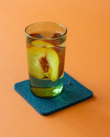Close-up of a green drinking glass filled with water and ice cubes with condensation moisture on a square Merino wool coaster in color Kombu