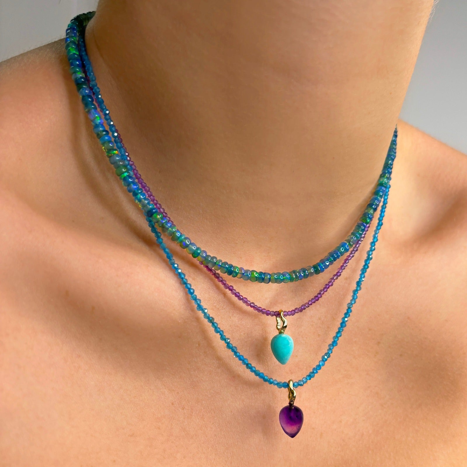 Two Shimmering slim gemstone beaded necklaces made of 2mm faceted stones in amethyst and Apatite on a gold linking lobster clasp.  Styled on a neck layered with two acorn drop charms and a denim opal necklace