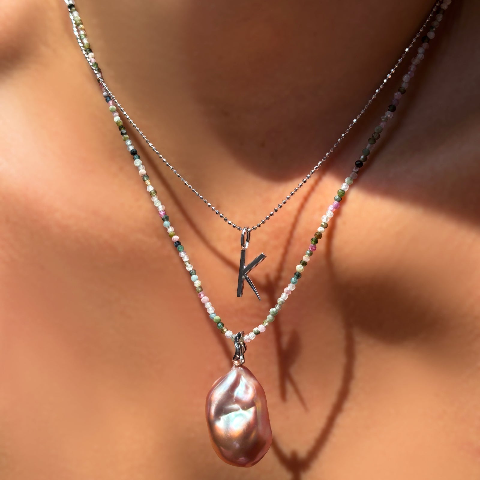 Layered necklace featuring a diamond-cut bead chain with a white gold 'K' letter charm and a tourmaline gemstone necklace with a blush baroque pearl charm