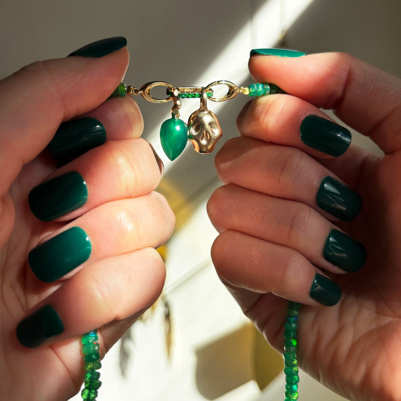 Green onyx drop charm, molten skull charm, small emerald charm lock, on a green faceted opal necklace in hand.