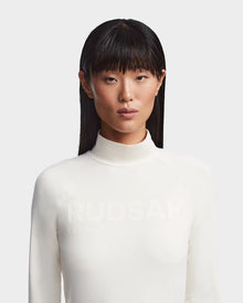 A woman with straight black hair and bangs wears the Rudsak KILTON - 8523533 Iceland fitted white mock-neck top featuring subtle branding, standing against a light grey background—a modern take on Canadian ski style.