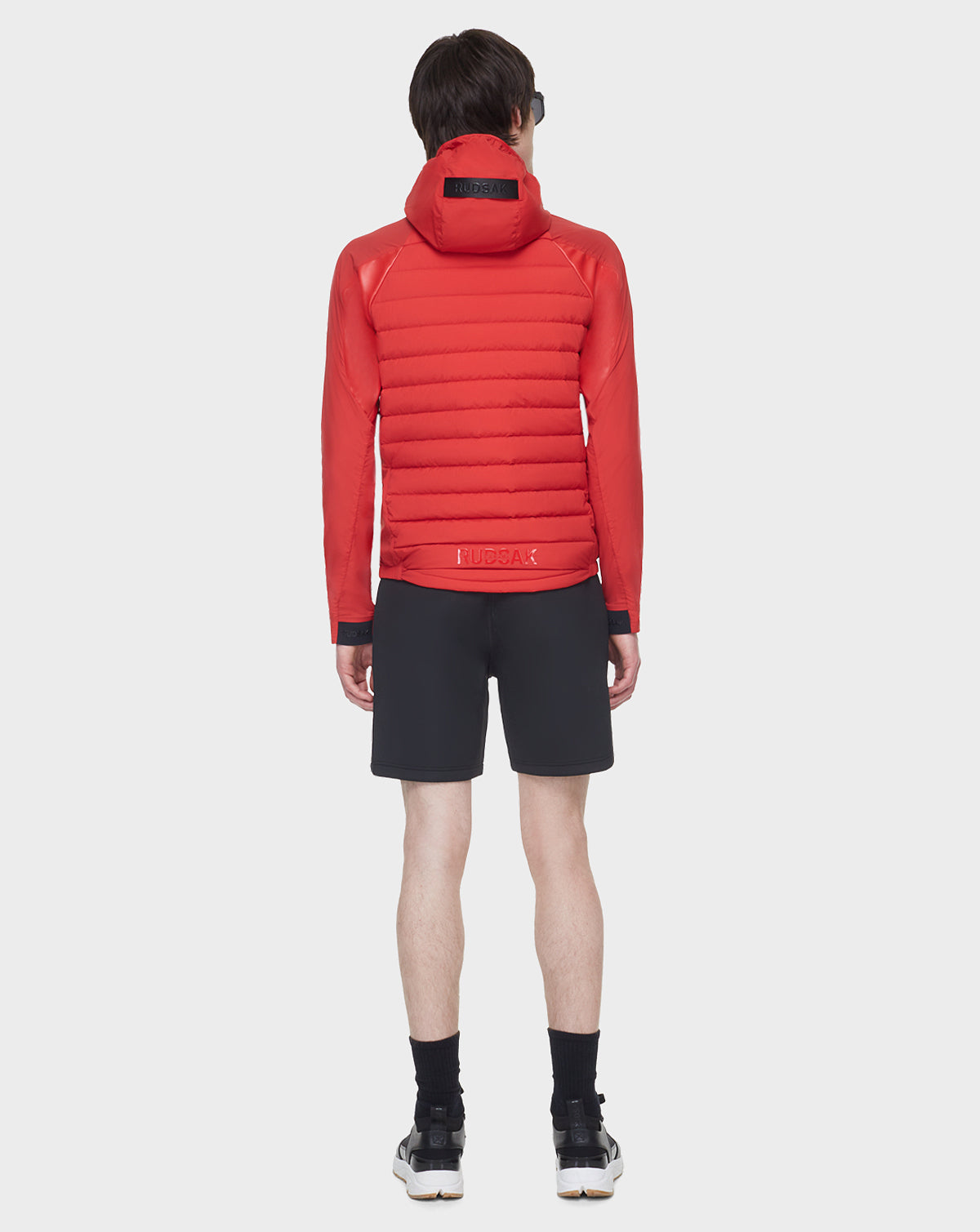 A person stands facing away against a white background, wearing the RUDSAK JARRY - 6122007 POPPY RED RDS-certified down puffer jacket with black shorts, socks, and shoes.