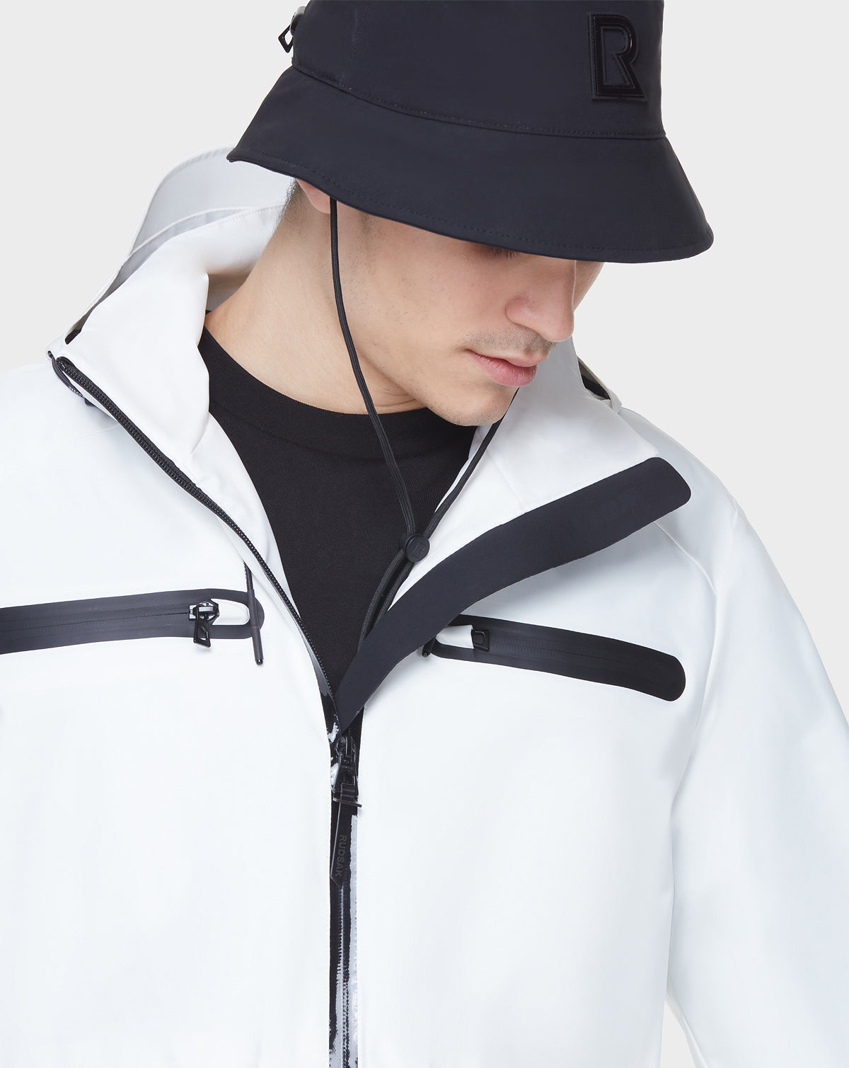A man wearing the RUDSAK JACY - 6122000 WHITE water-resistant trench coat and a black bucket hat looks down against a plain light gray background.