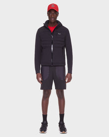 A man faces forward on a plain white background wearing the RUDSAK JAS - 6122006 BLACK men’s windbreaker with RDS-certified down, paired with a red cap, red shirt, black shorts with gray pockets, black socks, and black sneakers.