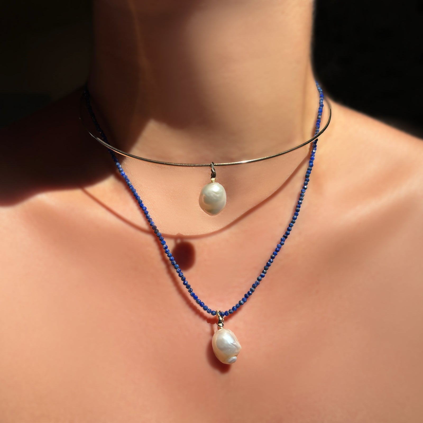 A layered look of a mini baroque pearl charm on a gold wire choker over a lapis slim gemstone necklace with a 2nd mini baroque pearl, shown on the neck in sunlight.