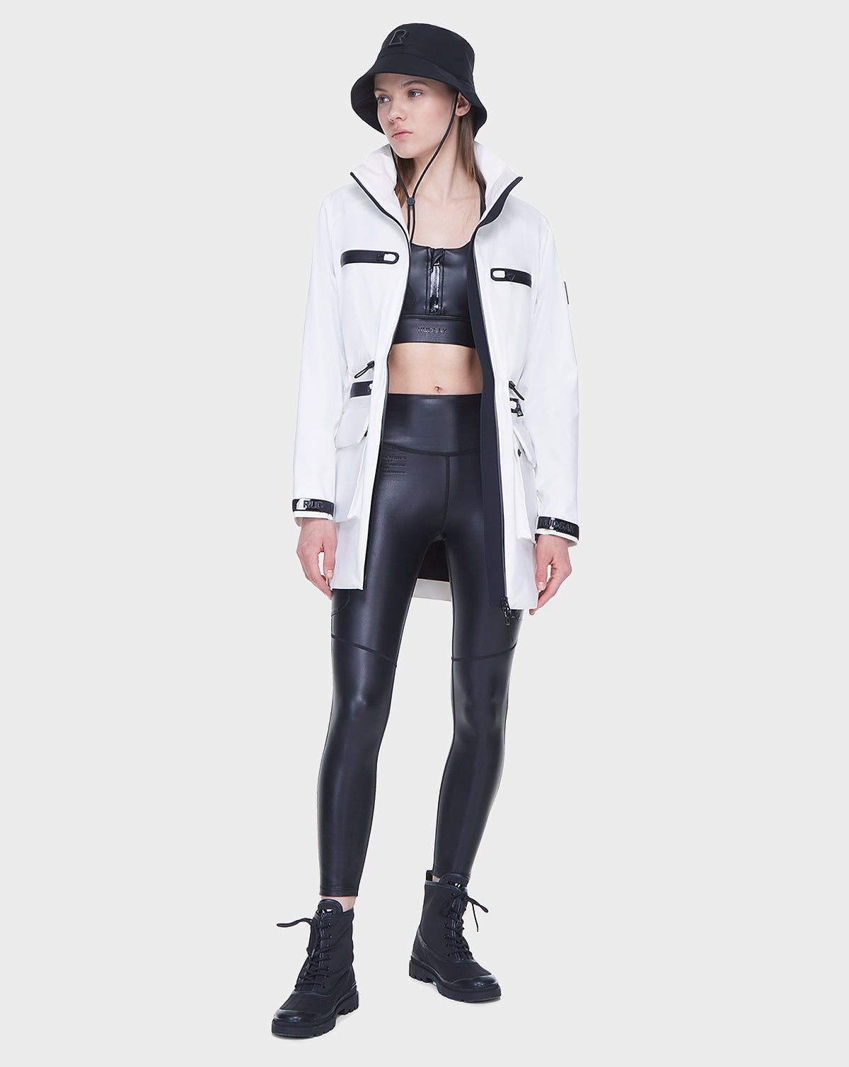 A woman stands against a plain light background wearing the Rudsak CAMY - 8122002 WHITE mid-length raincoat with black details, paired with a black bucket hat, crop top, high-waisted leggings, and boots.