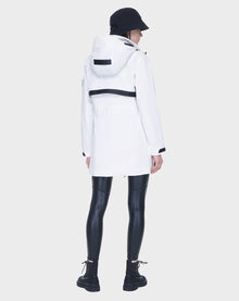 A person in the Rudsak CAMY - 8122002 WHITE water-resistant trench coat, black bucket hat, shiny black leggings, and black boots stands with their back to the camera against a plain light gray background.