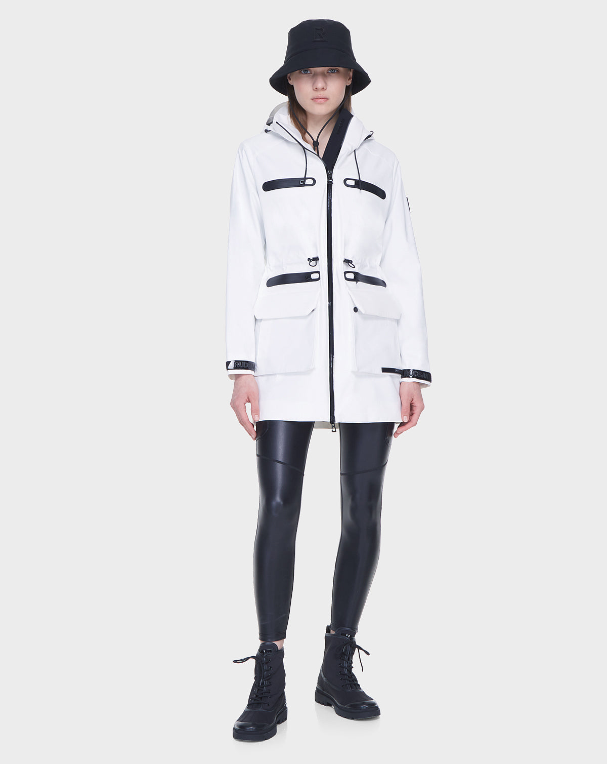 A person stands against a plain light gray background wearing the Rudsak CAMY - 8122002 WHITE water-resistant trench coat with black accents, paired with a black bucket hat, shiny black leggings, and lace-up boots.